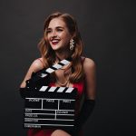 Lady with red lips holding clapperboard on black background. Happy woman in luxury silk dress smiling and posing on isolated backdrop