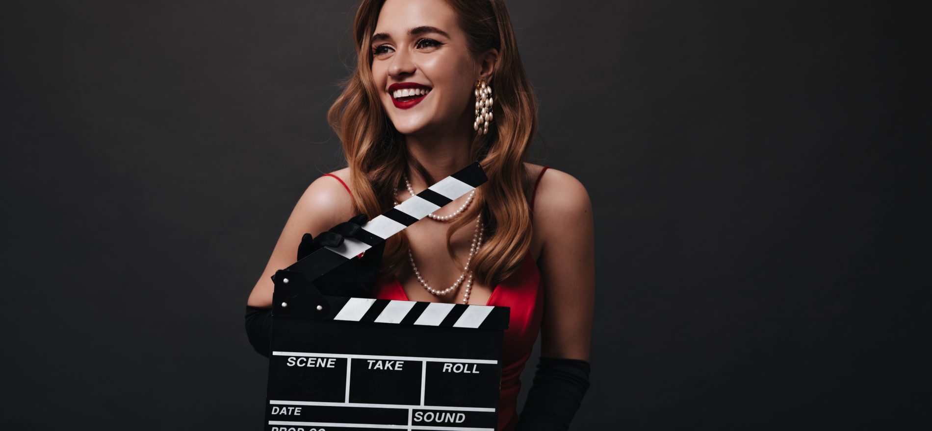 Lady with red lips holding clapperboard on black background. Happy woman in luxury silk dress smiling and posing on isolated backdrop