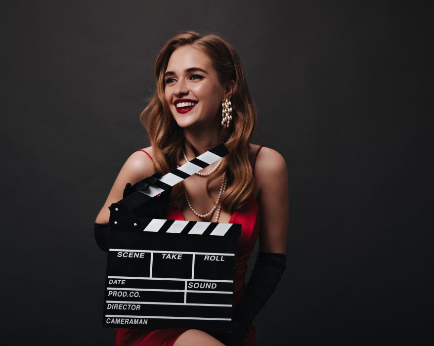 Lady with red lips holding clapperboard on black background. Happy woman in luxury silk dress smiling and posing on isolated backdrop