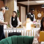 smiling concierge assists elderly male guest front desk while caucasian senior woman enjoys reading novel lounge area hotel lobby features comfy sofa reception counter reservations