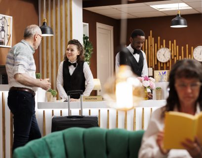 smiling concierge assists elderly male guest front desk while caucasian senior woman enjoys reading novel lounge area hotel lobby features comfy sofa reception counter reservations