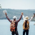 A mand and a women travelling together standing in front of a lake with their hands raised high