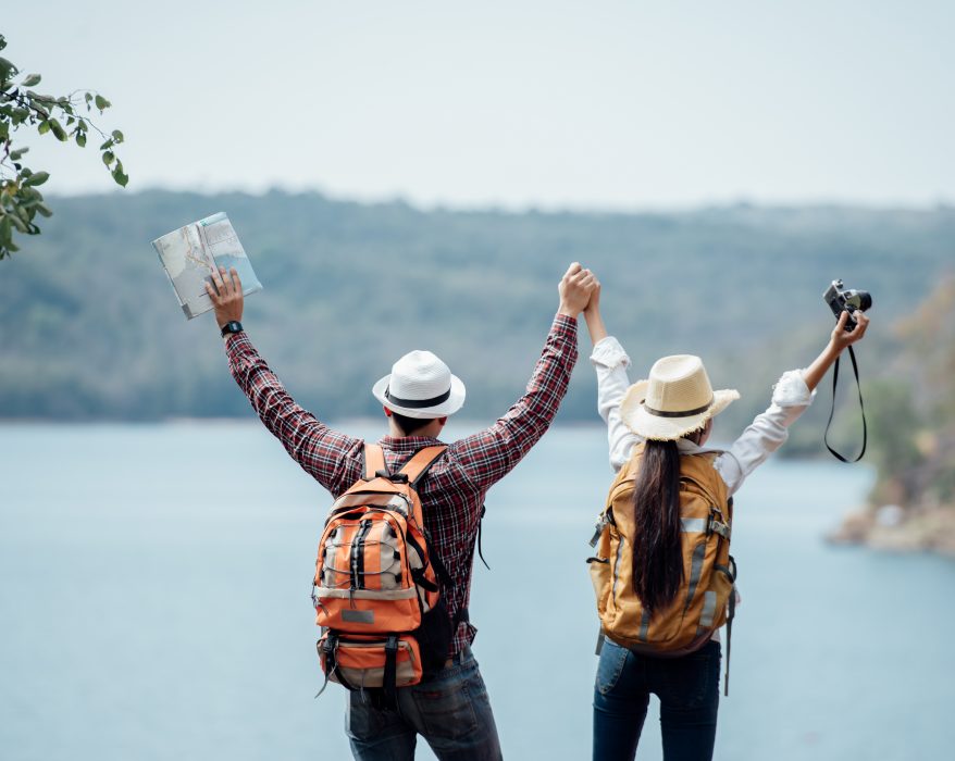 A mand and a women travelling together standing in front of a lake with their hands raised high