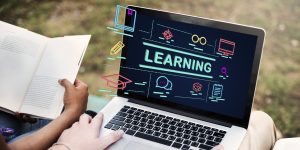 Student studying with a laptop showing online learning icons and the word “learning” on screen while reading a book outdoors, representing online education and digital courses.