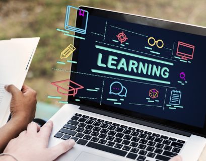 Student studying with a laptop showing online learning icons and the word “learning” on screen while reading a book outdoors, representing online education and digital courses.