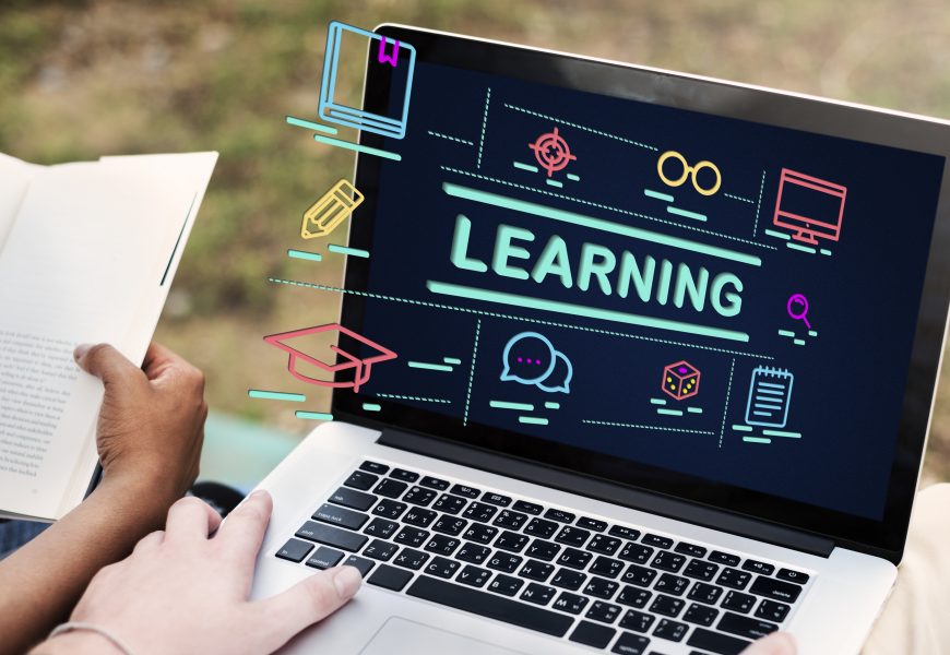 Student studying with a laptop showing online learning icons and the word “learning” on screen while reading a book outdoors, representing online education and digital courses.