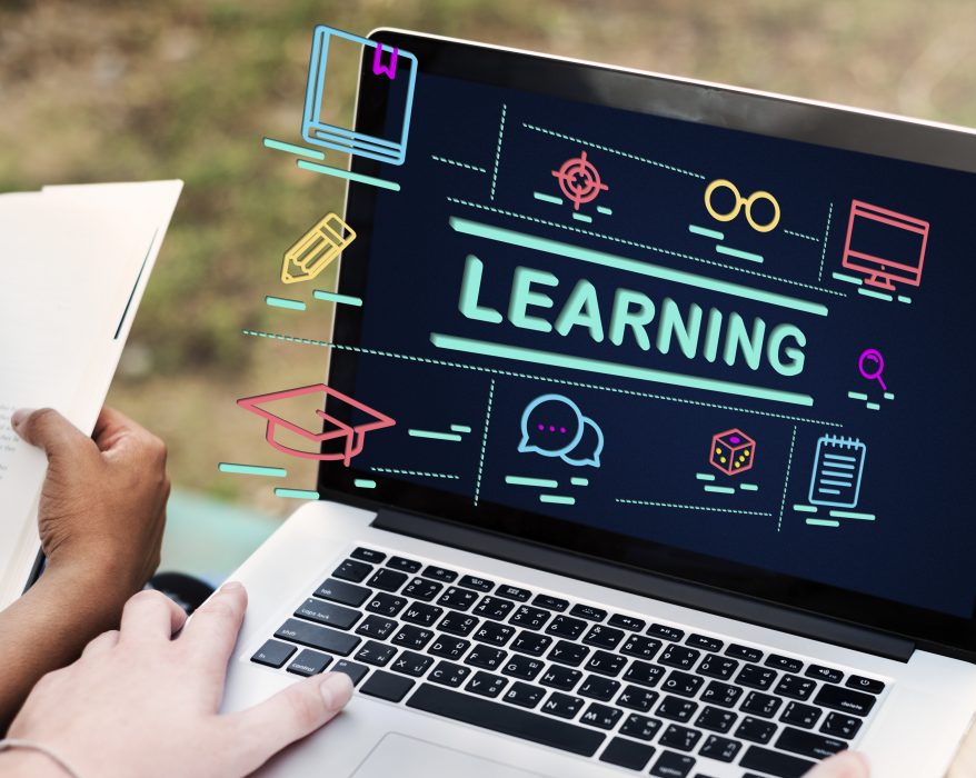 Student studying with a laptop showing online learning icons and the word “learning” on screen while reading a book outdoors, representing online education and digital courses.