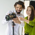 Two photographers smiling while adjusting a DSLR camera on a tripod in a photography studio, reviewing camera settings during a photoshoot. 📷