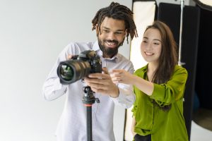 Two photographers smiling while adjusting a DSLR camera on a tripod in a photography studio, reviewing camera settings during a photoshoot. 📷