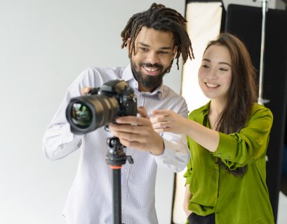 Two photographers smiling while adjusting a DSLR camera on a tripod in a photography studio, reviewing camera settings during a photoshoot. 📷