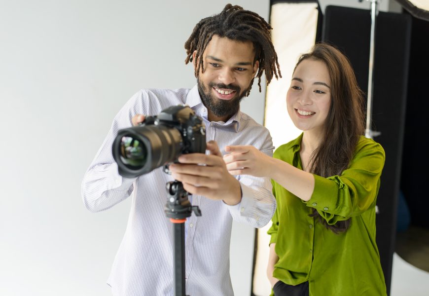 Two photographers smiling while adjusting a DSLR camera on a tripod in a photography studio, reviewing camera settings during a photoshoot. 📷
