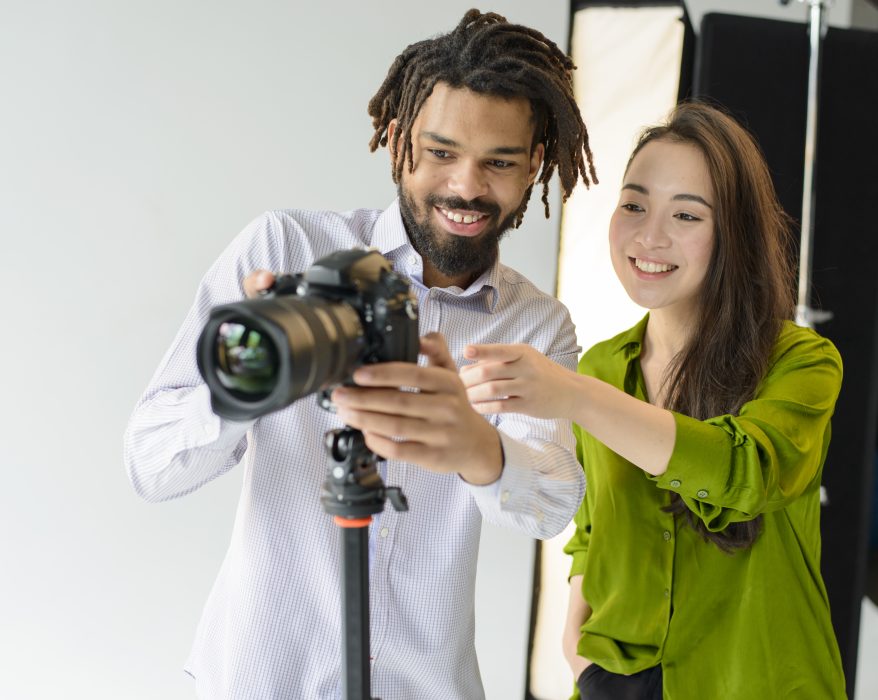 Two photographers smiling while adjusting a DSLR camera on a tripod in a photography studio, reviewing camera settings during a photoshoot. 📷